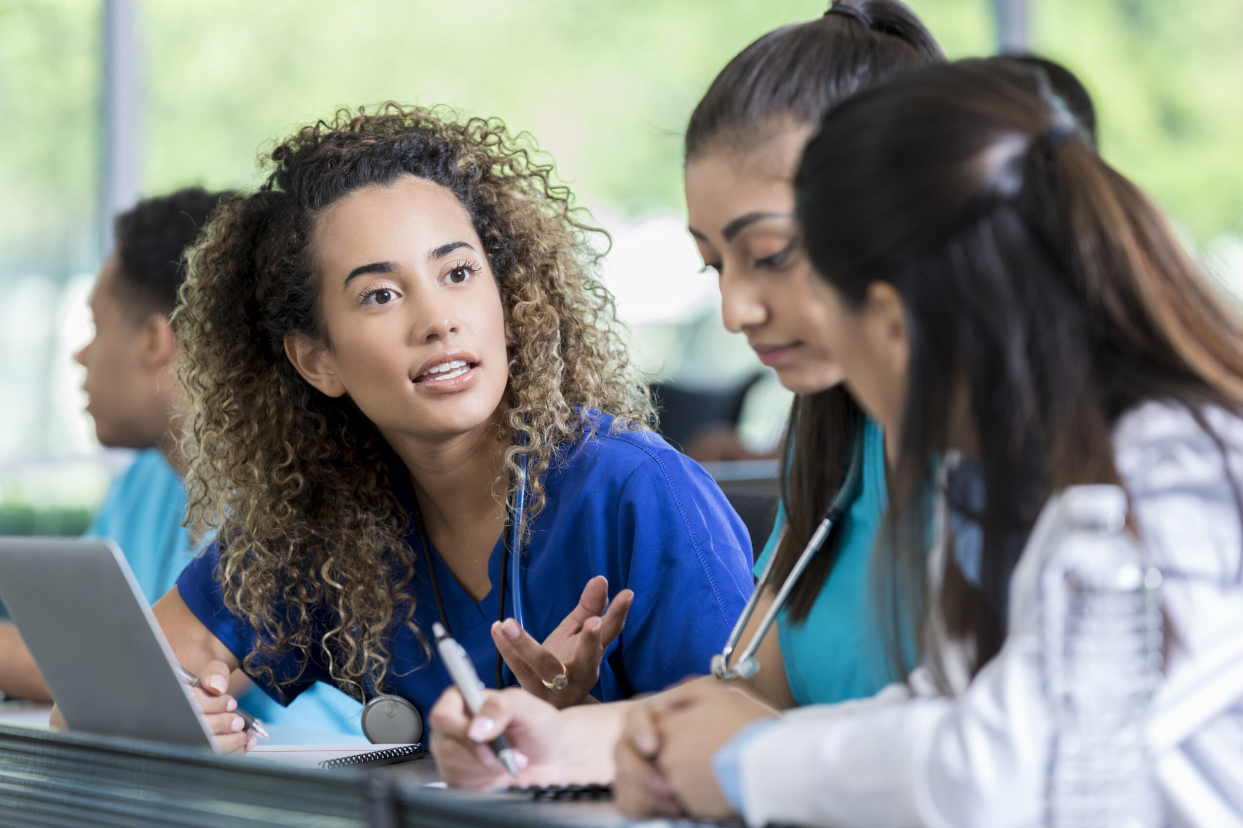 Three medical trainees sit in their classroom in front of a laptop and work on an assignment together.  One young student talks as the others listen and take notes.